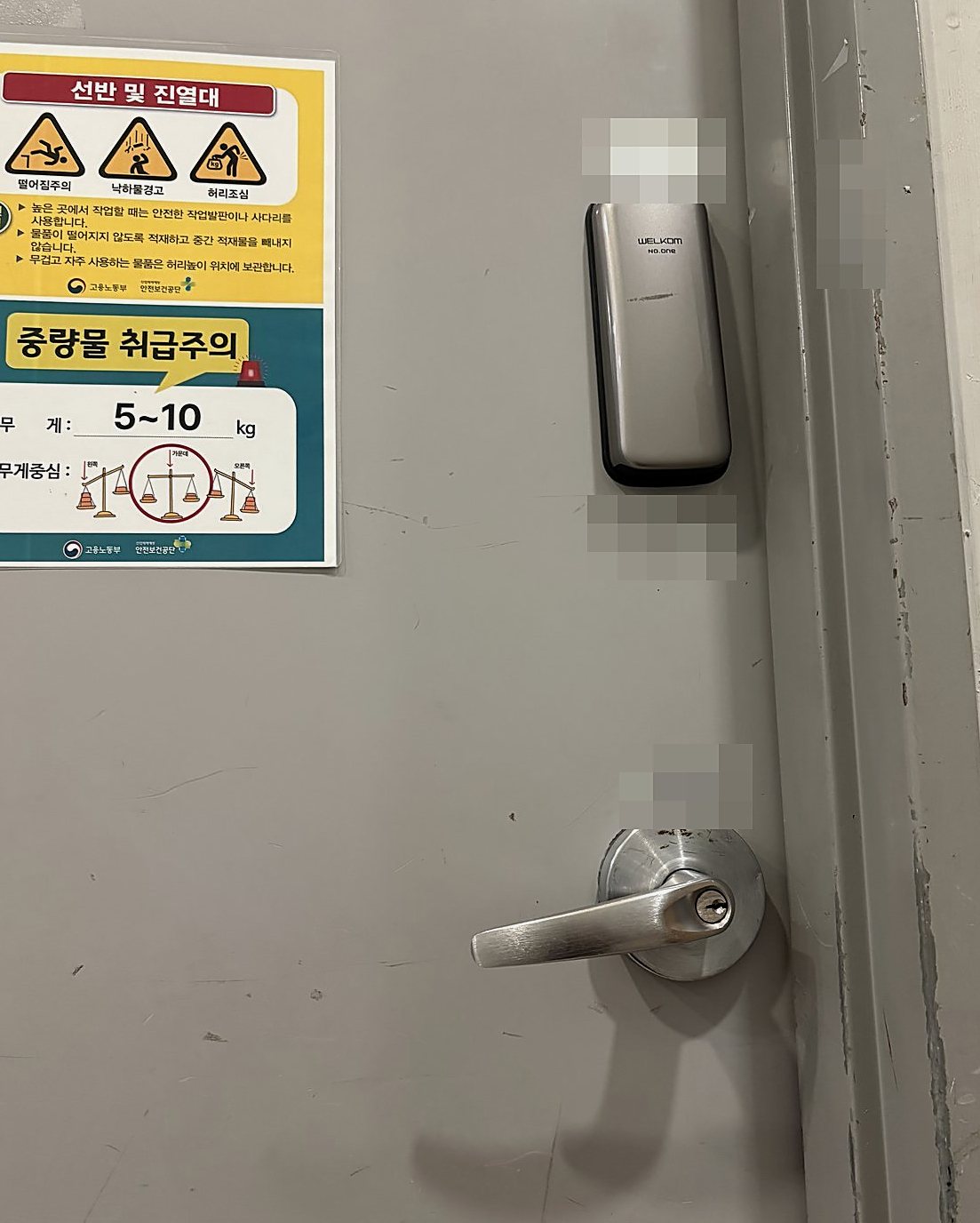 양천구 신월5동 출장 — 회색 철문 위쪽의 작은 은색 번호키와 아래쪽 열쇠 구멍이 있는 레버 손잡이, 왼쪽 안내 포스터